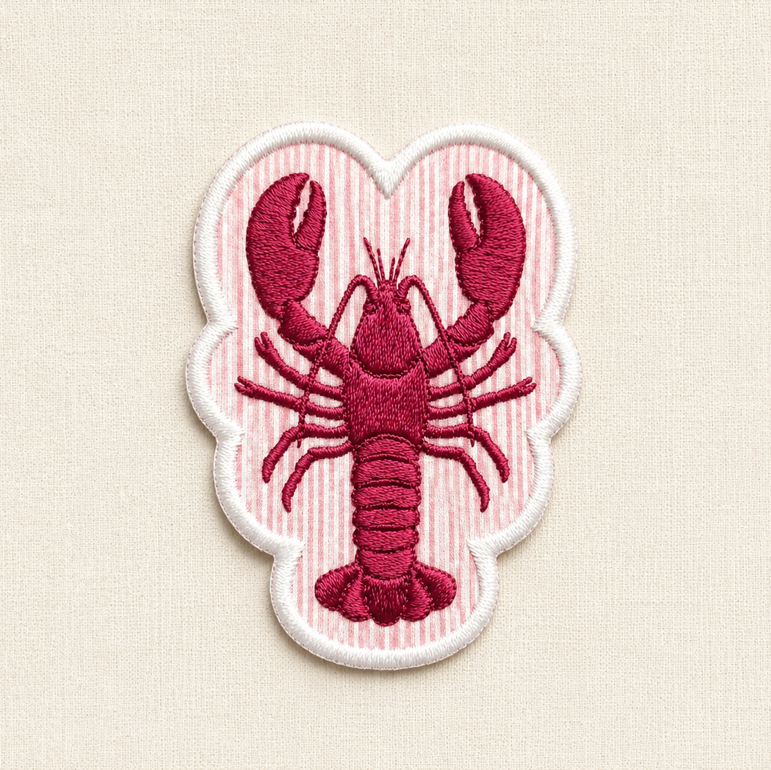 A close-up of a lobster shaped embroidered patch. In the center is a red lobster, set against a background of alternating horizontal stripes similar to seersucker. The entire patch is enclosed by a clean, raised white merrowed border.