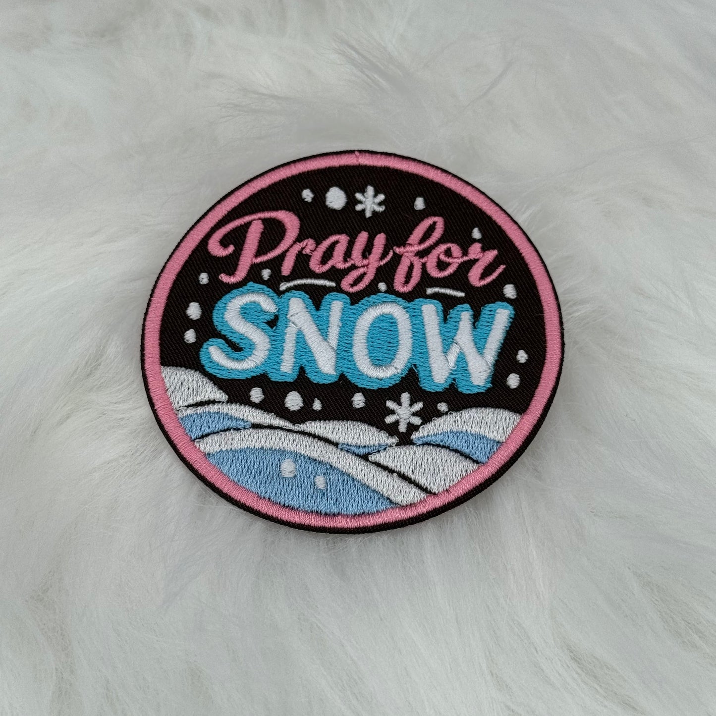 Round embroidered patch with 'Pray for Snow' text with soft mountains and snow in the background. Colors are pink, blue and white on a brown background. This is displayed on a fluffy white fur blanket. 

Great for winter and après ski clothing, bags, accessories and gifts! 