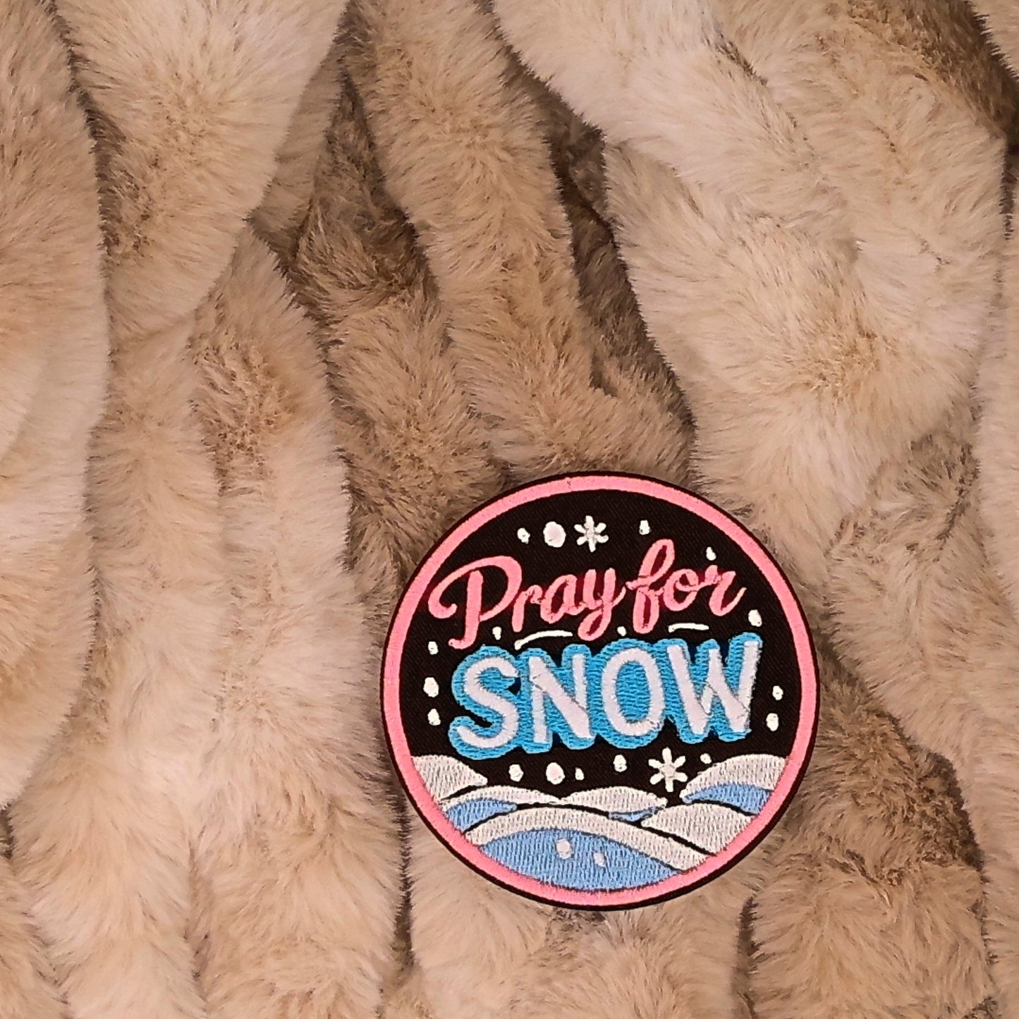 Round embroidered patch with 'Pray for Snow' text with soft mountains and snow in the background. Colors are pink, blue and white on a brown background. This is displayed on a fluffy brown fur blanket. 

Great for winter and après ski clothing, bags, accessories and gifts! 