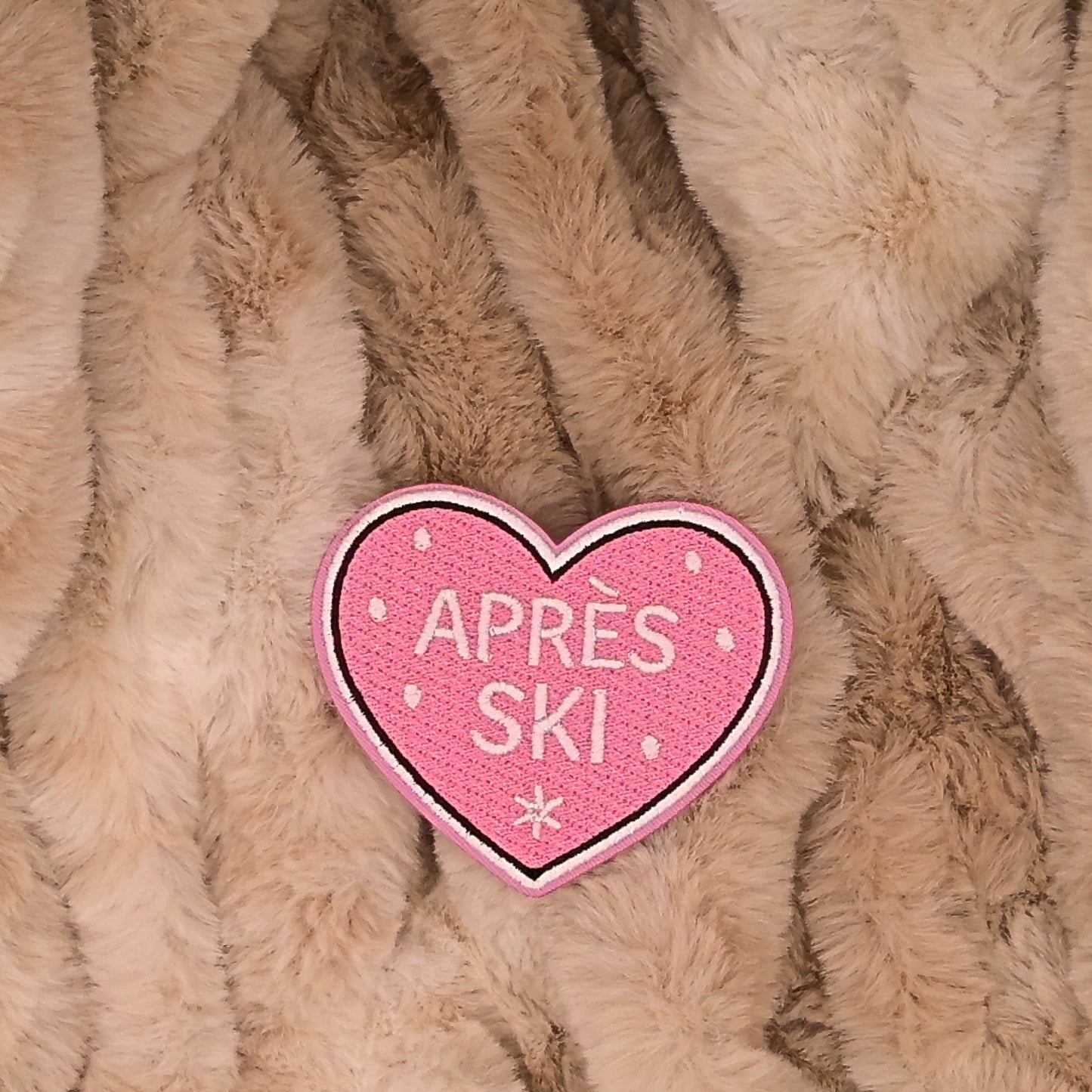 Heart Shaped embroidered patch with 'Apèrs ski' text with snow in the background. Colors are pink, and white on a brown background. This is displayed on a fluffy brown fur blanket. 

Great for winter and après ski clothing, bags, accessories and gifts! 