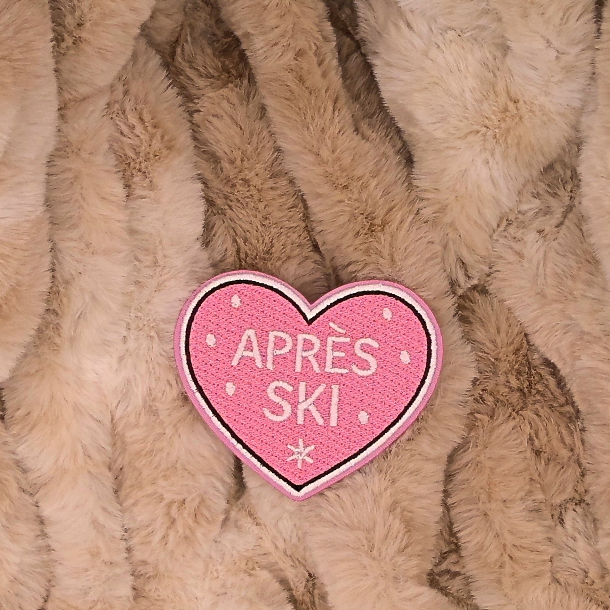 Heart Shaped embroidered patch with 'Apèrs ski' text with snow in the background. Colors are pink, and white on a brown background. This is displayed on a fluffy brown fur blanket. 

Great for winter and après ski clothing, bags, accessories and gifts! 